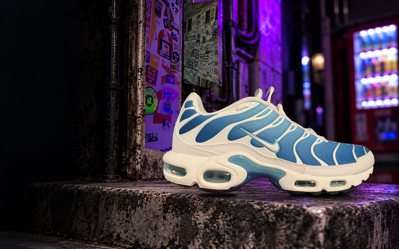 Blue-and-white Nike Air Max Plus TN on a concrete ledge in a purple-lit alley.